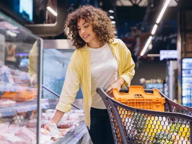 Kobieta z wózkiem na zakupy przegląda produkty w lodówce sklepowej.