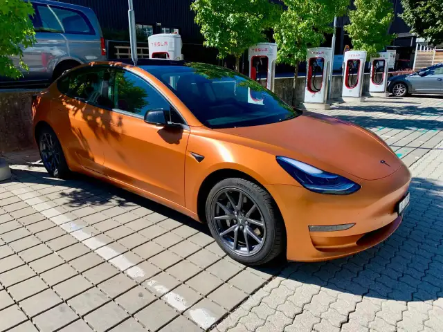 Pomarańczowa Tesla na tle ładowarek Supercharger.
