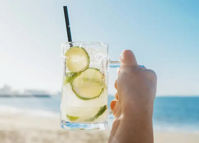 Orzeźwiająca Caipirinha czy Caipiroska z limonką i lodem w dłoni na tle plaży i morza. Idealny napój na upalny dzień.