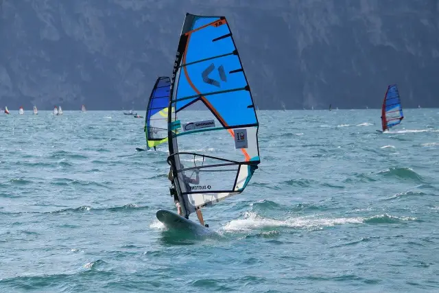 Osoba na desce windsurfingowej z niebieskim żaglem na wzburzonym morzu.