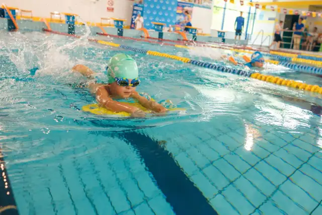 Chłopiec w okularach i czepku pływacki ćwiczy w basenie z deską. To świetny sposób, jak nauczyć dziecko pływać, budując pewność siebie w wodzie.