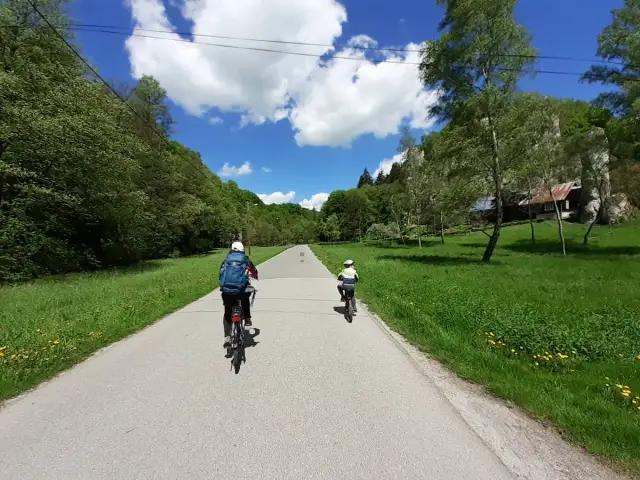 Dwie osoby na rowerach jadą malowniczą trasą rowerową w Ojcowie. W tle zielone wzgórza i błękitne niebo z chmurami.