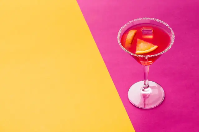 Elegancki cosmopolitan drink co zamiast żurawiny, z pomarańczą i lodem, na tle żółto-różowym.