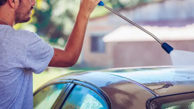 Mężczyzna pokazuje, jak myć auto na myjni, używając myjki ciśnieniowej do spłukiwania dachu samochodu.