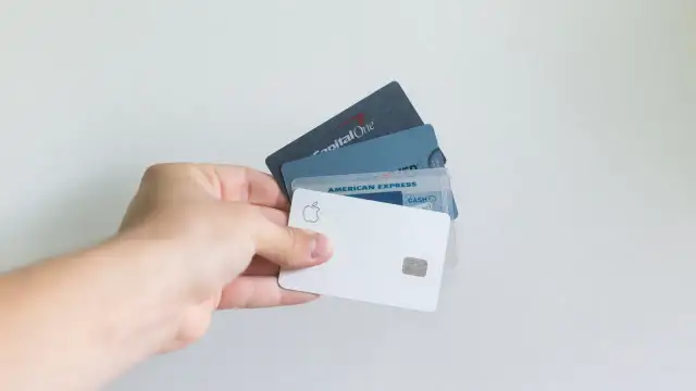 Dłoń trzyma kilka kart płatniczych, w tym Apple Card, American Express i Capital One. Standardowe wymiary karty bankomatowej są widoczne.