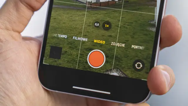 Dłoń trzyma iPhone'a z otwartą aplikacją aparatu, widoczny przycisk nagrywania wideo.