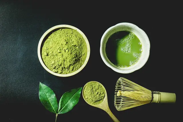 Miska z zielonym proszkiem matcha, filiżanka napoju, liście i bambusowa miotełka.