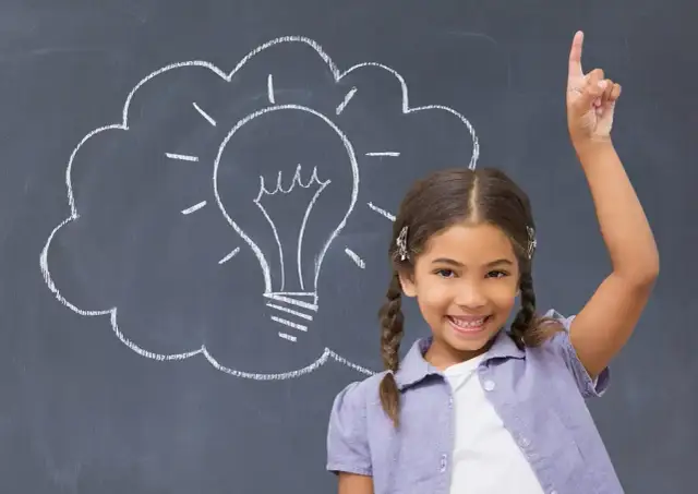 Uśmiechnięta dziewczynka z uniesionym palcem na tle tablicy z rysunkiem żarówki.