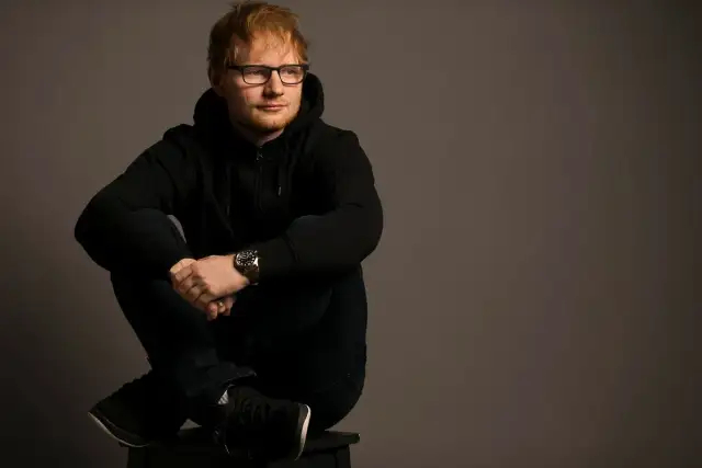 Ed Sheeran w czarnej bluzie z kapturem i okularach siedzi ze skrzyżowanymi nogami.