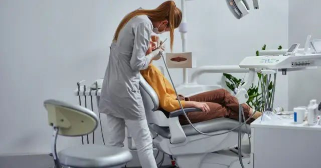 Dentystka w masce i rękawiczkach wykonuje zabieg pacjentowi siedzącemu na fotelu dentystycznym.