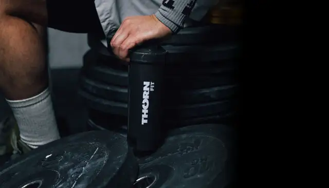 Dłoń trzyma czarny shaker z logo "THORN FIT" na tle stosu talerzy treningowych.