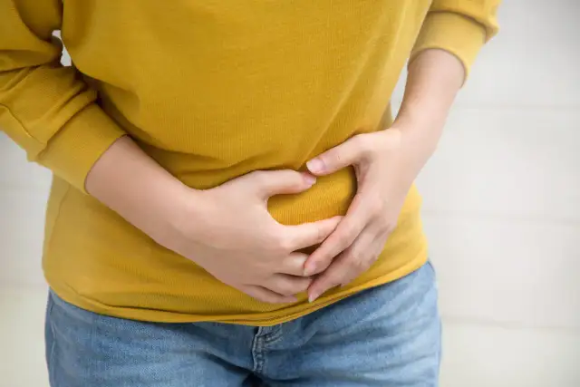 Osoba w żółtej bluzce trzyma się za brzuch, odczuwając ból. Szuka informacji, jak rozpuścić kamienie żółciowe.
