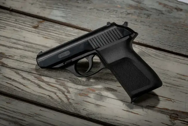 Czarny pistolet P.M.G.-23 leżący na zniszczonej drewnianej desce.
