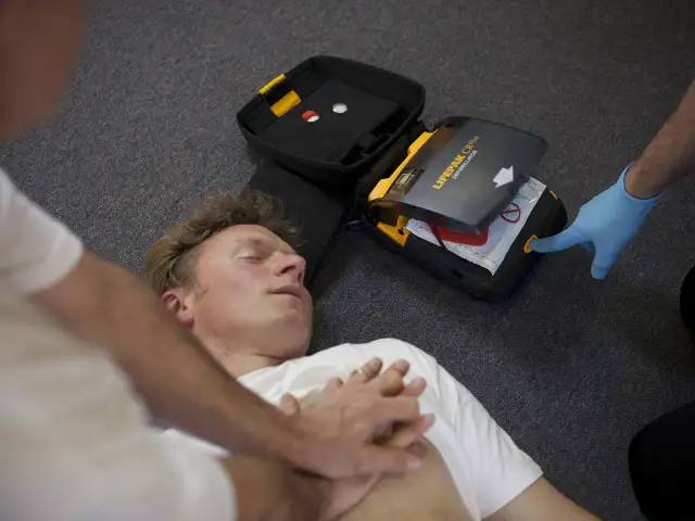 Osoba w niebieskiej rękawiczce naciska przycisk defibrylatora obok nieprzytomnego mężczyzny.