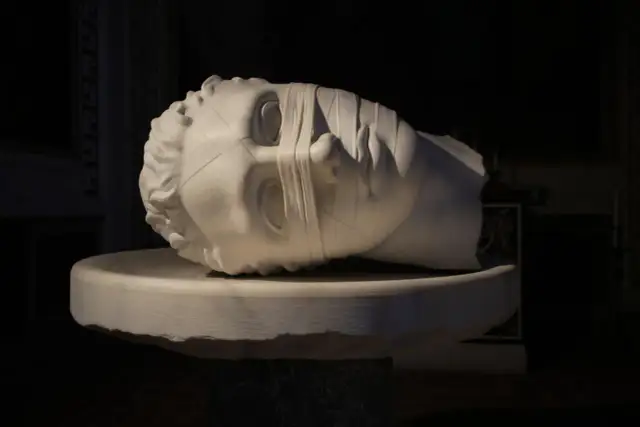 Biała marmurowa rzeźba głowy z opaską na oczach, leżąca na kamiennym postumencie.