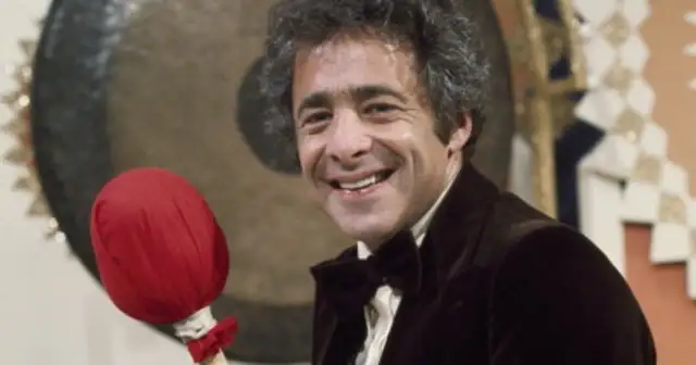 Uśmiechnięty mężczyzna w muszce i aksamitnej marynarce trzyma czerwony bębenek.