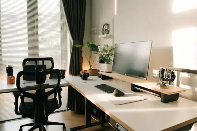 Nowoczesny gabinet w domu z biurkiem, monitorem, klawiaturą, myszką i wygodnym fotelem. Słońce wpada przez okno, tworząc przytulną atmosferę.