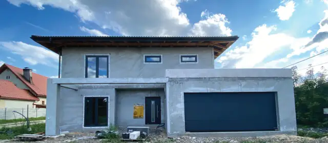 Budowa domu z szarym tynkiem, czarnymi oknami i bramą garażową pod błękitnym niebem.