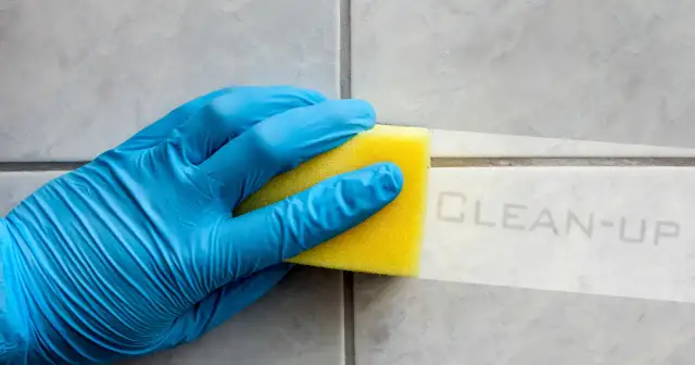 Ręka w niebieskiej rękawiczce czyści fugę żółtą gąbką.