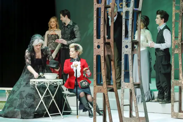 Scena z przedstawienia "Teatr kwadrat za jakie grzechy". Kobieta w czarnej sukni nalewa coś z misy, obok siedzi dama w czerwonym żakiecie.