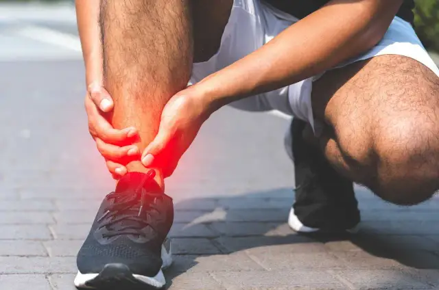 Osoba trzymająca się za bolesną, czerwoną kostkę w bucie sportowym.