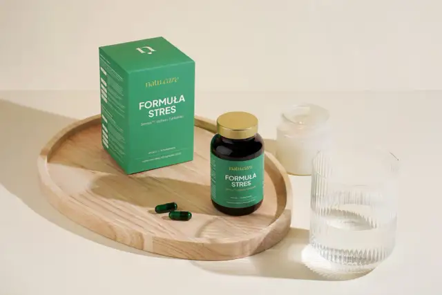 Zielone opakowanie i kapsułki suplementu diety "Formuła Stres" na drewnianej tacy, obok szklanki wody i świecy.
