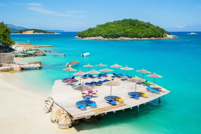 Rajska plaża z leżakami i parasolami, idealna na tanie kierunki na wakacje. Turkusowa woda, łodzie i zielona wyspa w tle.