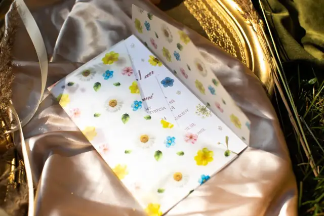 Otwarta koperta z zaproszeniem na ślub ozdobiona akwarelowymi kwiatami, leżąca na satynowej tkaninie.