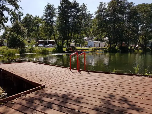 Drewniany pomost z czerwonymi poręczami nad jeziorem. W tle drzewa, budynki i samochody. Idealne miejsce na łagów camping.