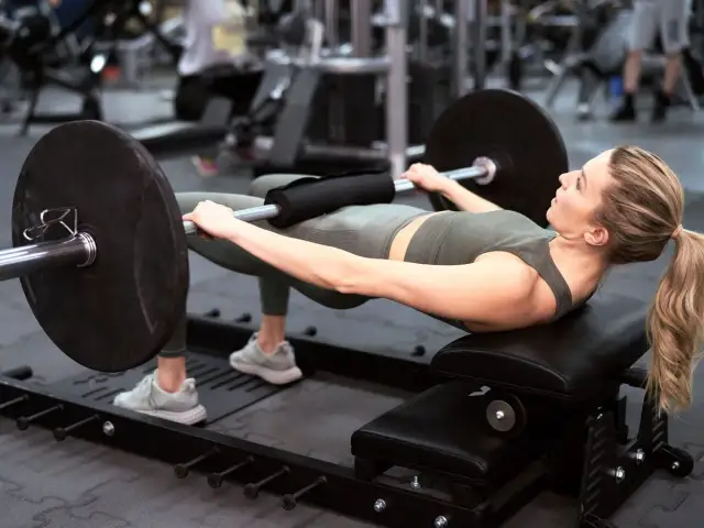 Kobieta wykonuje hip thrust z obciążeniem na ławce. Ćwiczenie to świetnie wzmacnia pośladki i jest możliwe do wykonania nawet w domu.