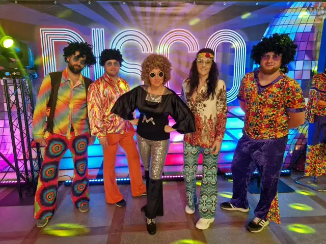 Pięć osób w kolorowych strojach disco z lat 70. na tle napisu "DISCO".