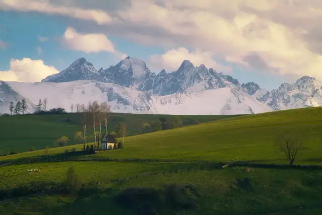 Zielone wzgórza, mały biały domek z drzewami i ośnieżone góry w tle.