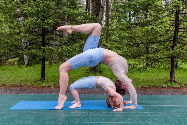 Dwie osoby wykonują ćwiczenia jogi w pozycji mostka na macie na tle lasu.