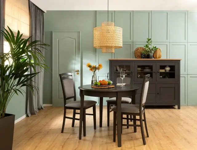 Jadalnia w odcieniu kolor szałwiowy. Okrągły stół z krzesłami, kredens i duża roślina tworzą przytulną atmosferę.
