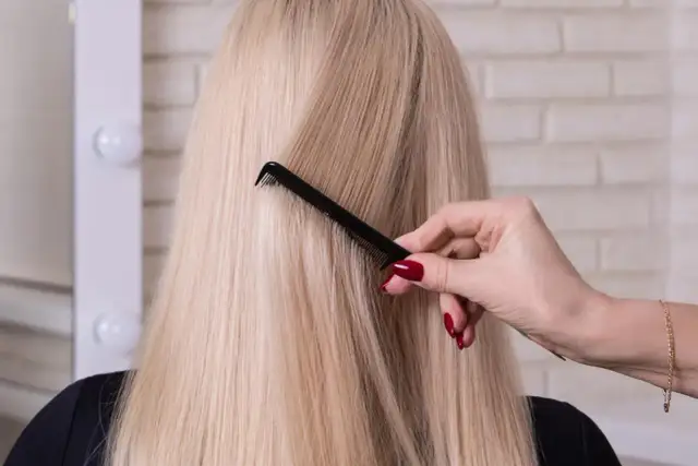 Dłoń z czerwonym lakierem do paznokci czesze długie, blond włosy. Zabieg botox na włosy rozjaśniane przywraca im blask.