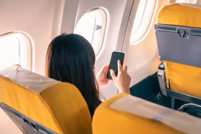 Kobieta w samolocie trzyma telefon, zastanawiając się, **co to tryb samolotowy**. Widok zza jej pleców na żółte siedzenia i okno.