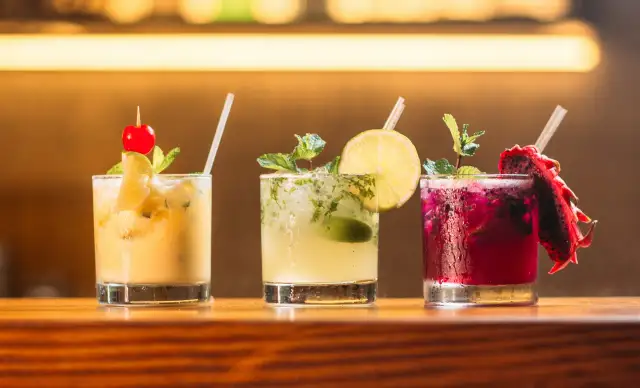Trzy kolorowe drinki na barze: żółty z wiśnią, zielony z limonką i miętą, oraz fioletowy z pitają. Idealne do letnich wieczorów, a może i z **kazbek peak brandy z czym pić**?