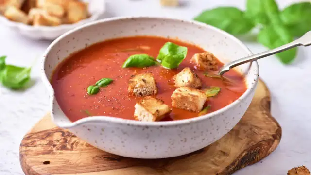 Miseczka zupy pomidorowej z grzankami i bazylią na drewnianej desce.