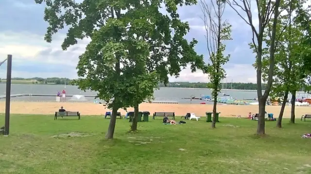 Zielone **łąka** nad **jeziorem**, piaszczysta plaża z ławkami i odpoczywającymi ludźmi, w tle łodzie i drzewa.