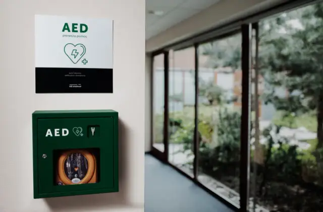 Zielona szafka z defibrylatorem AED na ścianie, obok tabliczka informacyjna.