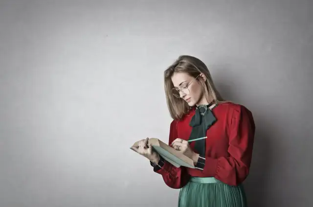 Młoda kobieta w okularach i czerwonej bluzce z zieloną kokardą pochyla się nad książką, notując coś ołówkiem. To inspiracja, jak napisać esej historyczny.