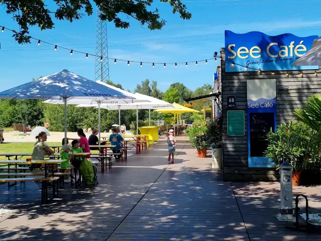 See Café mit Tischen und Gästen unter Sonnenschirmen am See.