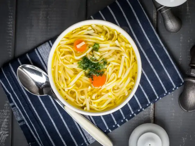 Miseczka zupy z makaronem, marchewką i natką pietruszki, obok łyżka i serwetka.