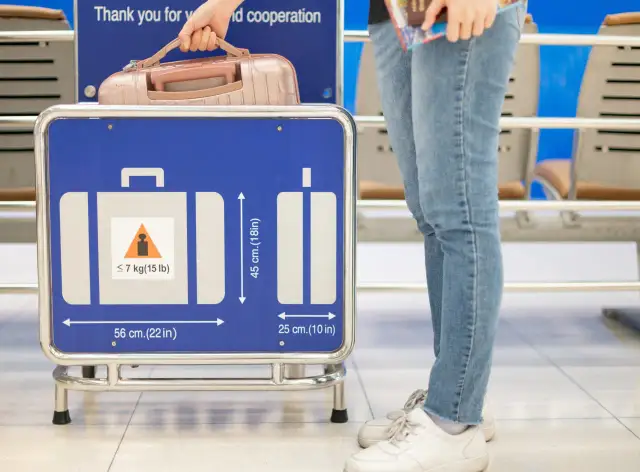 Person mit Handgepäck vor einem Schild mit Gepäckmaßen und Gewichtsbeschränkung.
