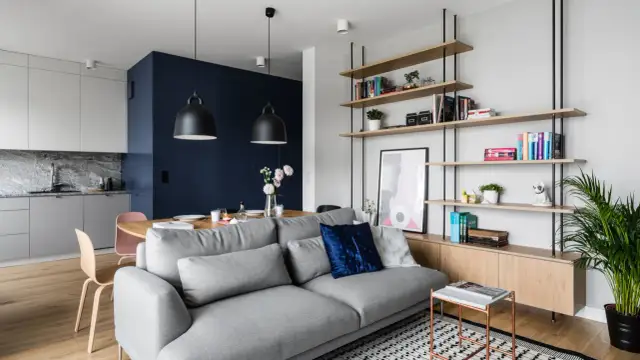 Szara sofa w małym salonie z półkami, stołem i aneksem kuchennym w tle.