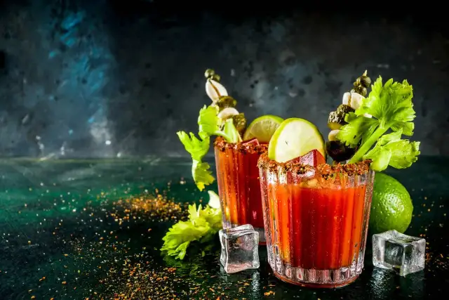 Dwa drinki Krwawa Mary z łodygami selera, limonką i piklami, ozdobione przyprawami i lodem.