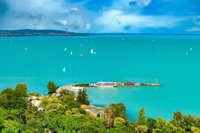 Turkusowe wody Balatonu na Węgrzech, pełne żaglówek i statków wycieczkowych, z zielonymi wzgórzami w tle.