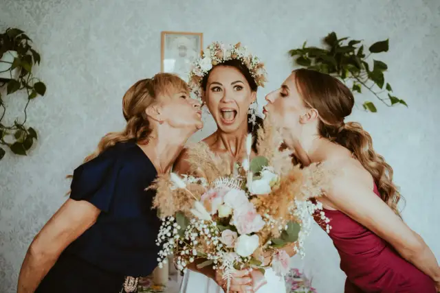 Panna młoda w wianku i dwie kobiety całujące ją w policzki, trzymają bukiet ślubny.