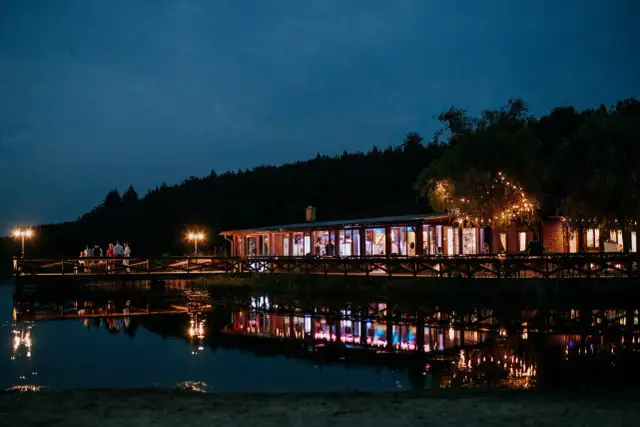 Wieczorny widok na budynek z przeszklonymi ścianami i oświetlonym molo nad jeziorem. Widać odbicia świateł w wodzie. To idealne miejsce na romantyczny wieczór w wodnym świecie Ostrzyce.