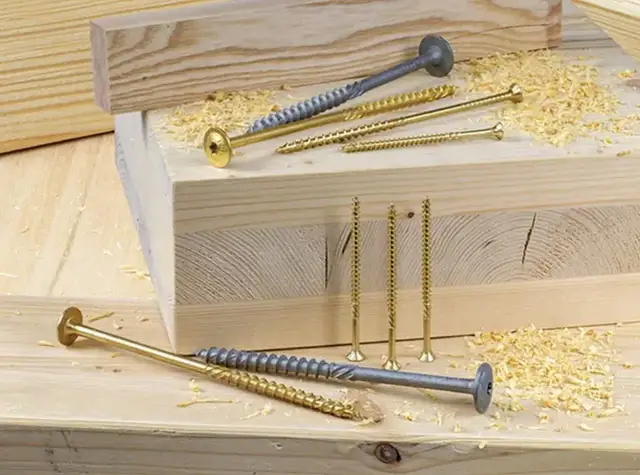 Wkręty do drewna różnej długości i koloru leżą na drewnianych deskach, wśród trocin.